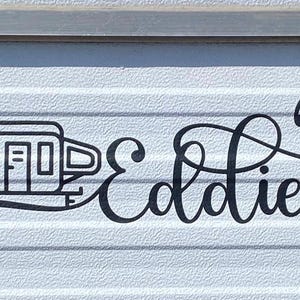 May include: Black and white illustration of a camper trailer with the word "Eldie" written in a cursive font.