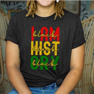 以下が含まれることがあります： 「I AM BLACK HISTORY」の文字が赤、黄、緑でプリントされた濃いグレーのTシャツ。「black」の文字は筆記体で書かれています。Tシャツはクルーネックで半袖です。