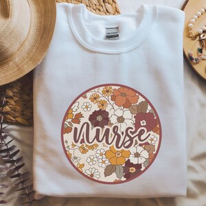 May include: White sweatshirt with a round floral design and the word "Nurse" in a brown script font.