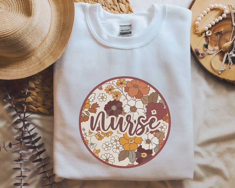May include: White sweatshirt with a round floral design and the word "Nurse" in a brown script font.