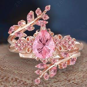 Pode incluir: Um anel de ouro rosa com uma gema rosa em forma de pêra cercada por gemas rosas menores. O anel apresenta um delicado design floral com folhas e flores.