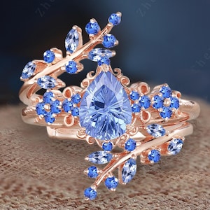May include: A rose gold ring with a pear-shaped blue gemstone surrounded by smaller blue gemstones. The ring features a delicate design of intertwined branches with leaves and flowers.