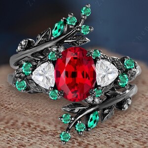 May include: A black metal ring featuring an oval-cut, vibrant red gemstone as the centerpiece, flanked by two white triangular stones. The ring is adorned with green and white accent stones and leaf-like details.