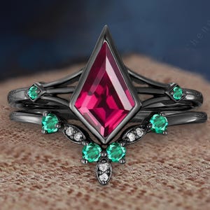 May include: A black engagement ring set with a large, kite-shaped, ruby-red gemstone. The set includes a matching band with emerald-green stones and small white accent stones. The rings are set against a neutral background.