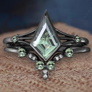 Op de afbeelding: Een zwarte verlovingsringset met een grote, vlieger-vormige, lichtgroene edelsteen. De set bevat een bijpassende band met kleinere groene stenen en delicate details. De ringen zijn geplaatst tegen een neutrale achtergrond.