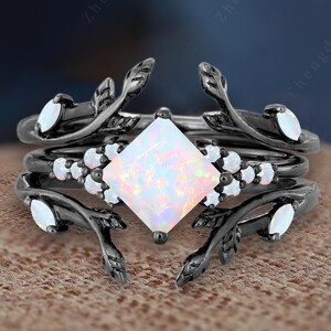May include: A three-piece black ring set. The rings feature a central square opal stone, surrounded by smaller white stones and leaf-like designs. The rings are set on a textured brown surface.