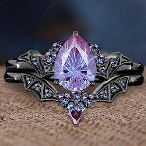 Może przedstawiać: Czarny zestaw pierścionków ślubnych z centralnym elementem w postaci fioletowego kamienia szlachetnego w kształcie gruszki. Pierścionek jest ozdobiony mniejszymi, okrągłymi i gruszkowatymi kamieniami szlachetnymi w odcieniach fioletu i błękitu. Obrączka ma dekoracyjny wzór.