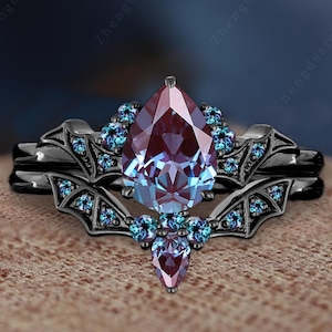 Może przedstawiać: Czarny zestaw pierścionków zaręczynowych z gruszkowym, zmieniającym kolor kamieniem szlachetnym. Pierścionek jest ozdobiony małymi, okrągłymi, niebieskimi kamieniami szlachetnymi. Opaska ma dekoracyjny wzór. Zestaw zawiera pasującą opaskę.