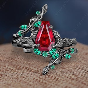Könnte beinhalten: Ein schwarzer und grüner Verlobungsring-Set mit einem großen roten Edelstein. Der Ring hat ein zartes Rankendesign mit Blättern und kleinen grünen Edelsteinen.