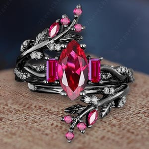 Puede incluir: Un conjunto de anillos negros con una gran gema roja en forma de marquesa y dos gemas rojas rectangulares más pequeñas. El anillo presenta un diseño floral con pequeñas gemas rosas y piedras de acento transparentes.