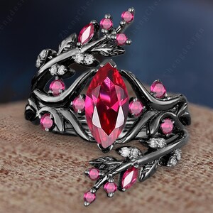 May include: Black ring with a large, marquise-cut, ruby-red gemstone as the centerpiece. The ring features a floral design with smaller pink gemstones and white accents. The ring is set against a neutral background.