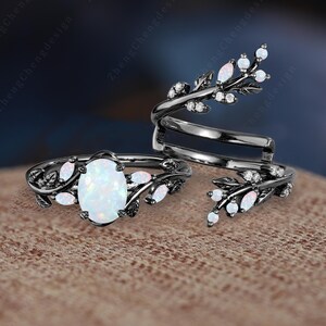 May include: Two black rings with white opal stones and leaf designs. The larger ring has a larger oval-shaped opal stone.