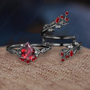 Puede incluir: Dos anillos negros con piedras preciosas rojas y blancas. El anillo más grande presenta una piedra roja en forma de pera rodeada de piedras blancas más pequeñas y hojas negras. El anillo más pequeño presenta un diseño de hoja negra con piedras rojas.