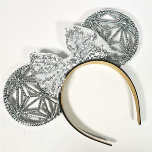 Könnte beinhalten: Silbernes Pailletten-Schleifen- und Ohren-Stirnband. Die Ohren haben ein geometrisches Design, und die Schleife ist mit Pailletten bedeckt. Das Stirnband ist außen schwarz und innen beige. Ideal für eine Themenparty oder Veranstaltung.