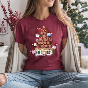 Könnte beinhalten: Burgunderfarbenes T-Shirt mit einem Weihnachtsbaum-Design mit Cartoon-Charakteren mit Weihnachtsmützen. Die Namen der BTS-Bandmitglieder sind in das Baumdesign integriert. Im Hintergrund befindet sich ein Schild mit der Aufschrift "Milk & Cookies".