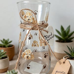 May include: A clear glass vase decorated with painted golden poodle faces and brown paw prints. The neck is wrapped in brown twine with a white heart-shaped ceramic tag reading "Hand painted" and a brown paper tag labelled "poodle" attached.