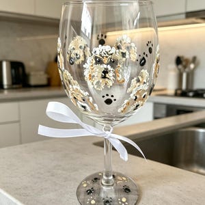 May include: A clear glass wine glass featuring hand-painted illustrations of small, fluffy, light-coloured dogs with black noses and eyes. The design includes scattered black paw prints around the dogs and on the base. A white ribbon is tied around the stem.