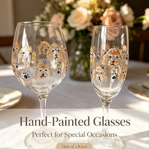 May include: Two clear glass vessels, a wide-bowled wine glass and a tall champagne flute, both featuring hand-painted gold and black illustrations of dog faces and paw prints encircling the exterior surface of the glass bowls.