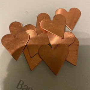 Puede incluir: Un montón de piezas de metal en forma de corazón de cobre. Los corazones están dispuestos en un patrón aleatorio sobre una superficie blanca.