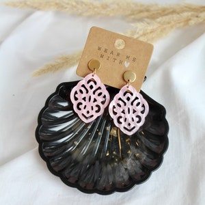 May include: Pink and gold earrings with a delicate, intricate design. The earrings are displayed on a black seashell-shaped dish with the text "Wear Me With" on a brown card.