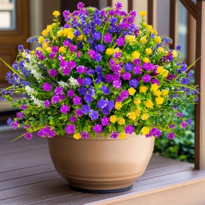 May include: A large, round, beige flower pot overflowing with colorful artificial flowers. The flowers are a mix of purple, yellow, and blue, with green foliage. The pot sits on a wooden surface, creating a vibrant outdoor display.