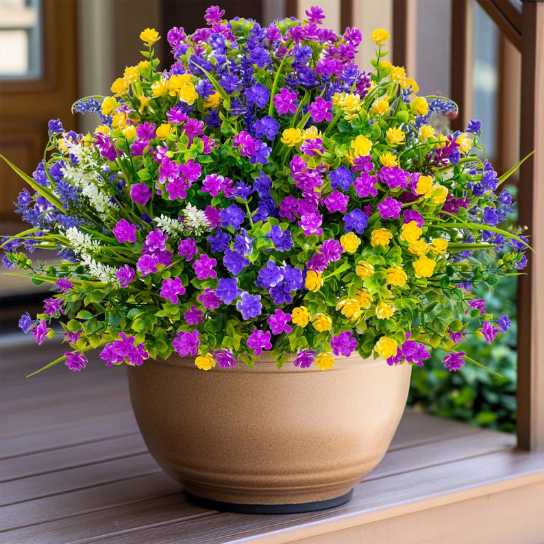 May include: A large, round, beige flower pot overflowing with colorful artificial flowers. The flowers are a mix of purple, yellow, and blue, with green foliage. The pot sits on a wooden surface, creating a vibrant outdoor display.