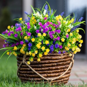 May include: A woven brown basket filled with colorful artificial flowers. The arrangement includes yellow, purple, blue, and white blossoms with green foliage. The basket has a rope handle and sits on green grass.