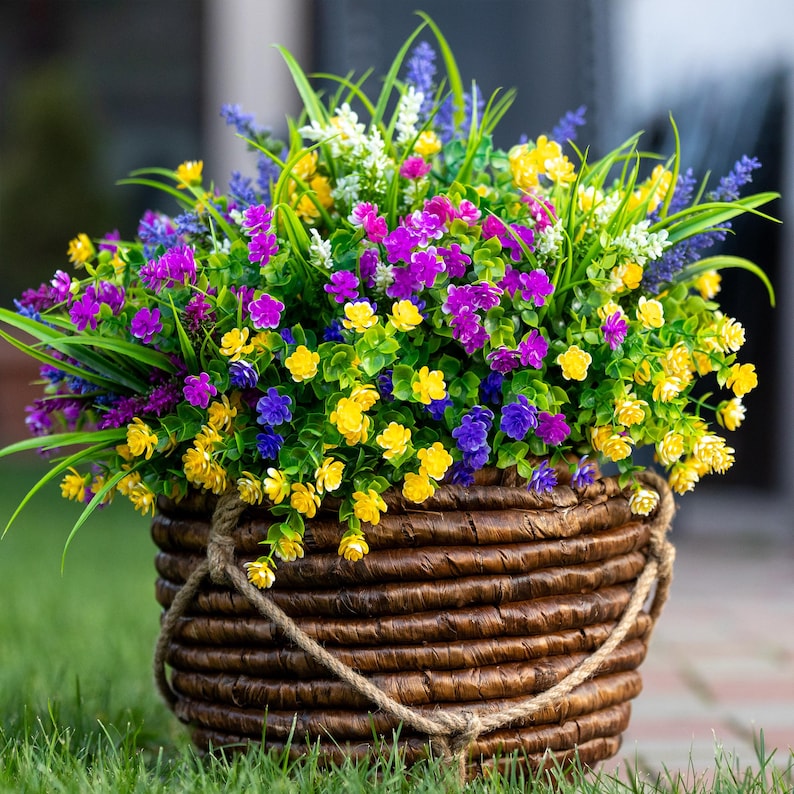 May include: A woven brown basket filled with colorful artificial flowers. The arrangement includes yellow, purple, blue, and white blossoms with green foliage. The basket has a rope handle and sits on green grass.