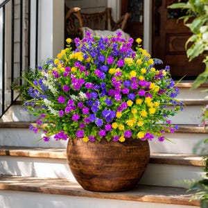May include: A large wooden planter filled with vibrant, colorful faux flowers. The arrangement features a mix of purple, yellow, and blue blossoms with green foliage. The planter sits on a wooden porch step, creating a welcoming outdoor display.