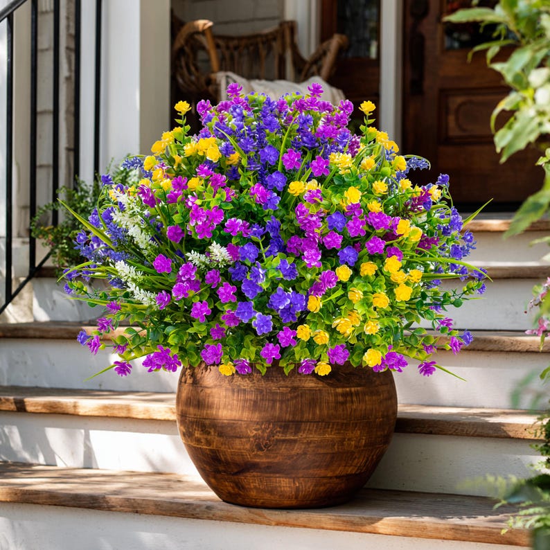 May include: A large wooden planter filled with vibrant, colorful faux flowers. The arrangement features a mix of purple, yellow, and blue blossoms with green foliage. The planter sits on a wooden porch step, creating a welcoming outdoor display.