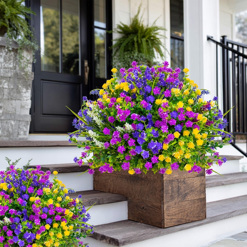 May include: A wooden planter box filled with vibrant artificial flowers in shades of purple, yellow, and blue. The flowers are arranged in a dense, bushy display, creating a colorful accent on a porch. A second planter box with similar flowers is also visible.