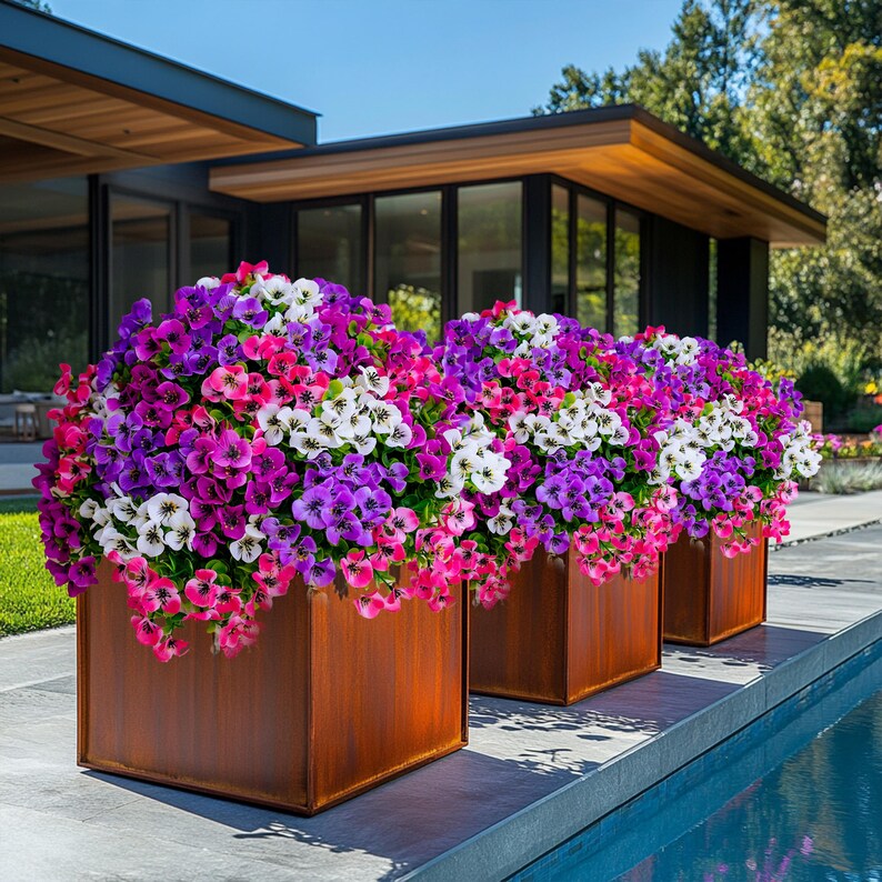 May include: Three square planters with a rusty brown finish are filled with a mix of pink, purple, and white flowers. The planters are arranged in a row on a stone patio next to a swimming pool.