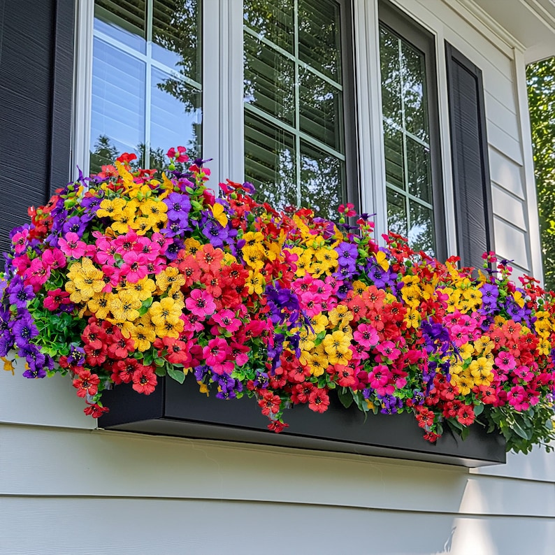 May include: A vibrant window box overflowing with colorful artificial flowers. The arrangement features a mix of red, pink, yellow, orange, and purple blooms, creating a cheerful display. The flowers are set in a black rectangular planter, adding a contrast to the bright colors.