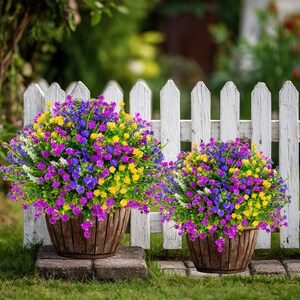 May include: Two wooden planters filled with vibrant, colorful flowers. The flowers are a mix of purple, yellow, blue, and white, creating a cheerful display. The planters sit on stone blocks in front of a white picket fence.