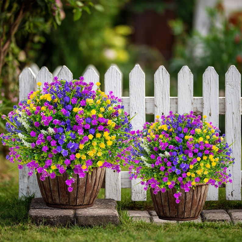 May include: Two wooden planters filled with vibrant, colorful flowers. The flowers are a mix of purple, yellow, blue, and white, creating a cheerful display. The planters sit on stone blocks in front of a white picket fence.