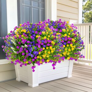 May include: A white rectangular planter box overflowing with colorful artificial flowers. The flowers are a mix of purple, yellow, blue, and white, creating a vibrant display. The planter is made of wood and sits on a wooden deck.