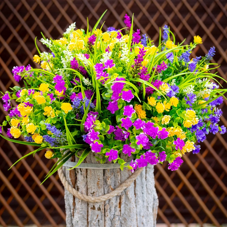 May include: A vibrant floral arrangement in a rustic wooden planter. The bouquet features a mix of yellow, purple, and blue flowers with green foliage. A rope handle adds a decorative touch to the planter, creating a natural aesthetic.