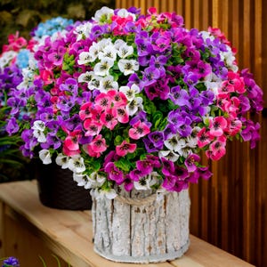 May include: A bouquet of purple, pink, and white flowers in a white planter with a wood-like texture. The planter is sitting on a wooden surface.