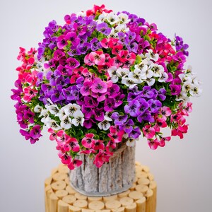 May include: A bouquet of artificial flowers in a white planter with a wood grain texture. The flowers are pink, purple, and white.