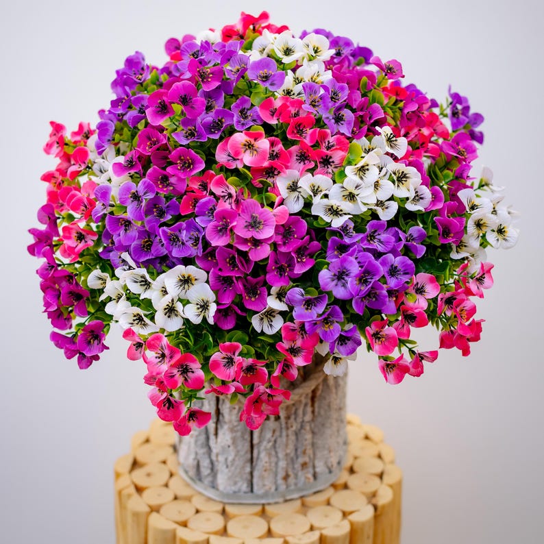 May include: A bouquet of artificial flowers in a white planter with a wood grain texture. The flowers are pink, purple, and white.