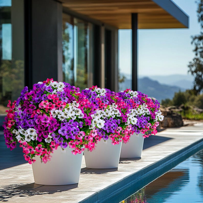 May include: Three white planters filled with colorful artificial flowers. The flowers are a mix of pink, purple, and white. The planters are sitting on a patio next to a swimming pool.