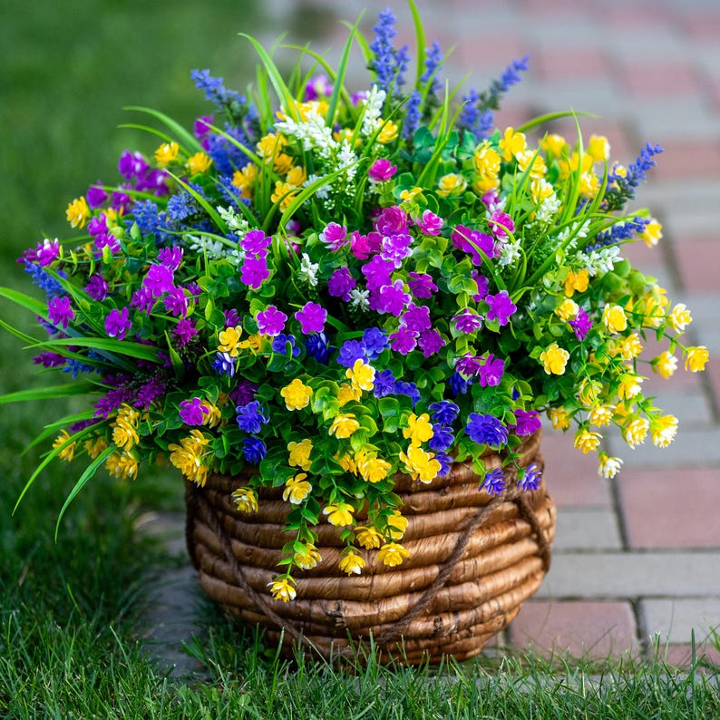 May include: A woven basket overflowing with colorful artificial flowers. The arrangement includes vibrant yellow, purple, blue, and white blossoms, with green foliage. The basket is made of brown woven material and sits on green grass.