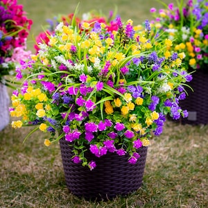 May include: A vibrant arrangement of artificial flowers in a dark brown woven planter. The colorful mix includes yellow, purple, pink, and white blossoms with green foliage. The planter sits on a grassy surface, showcasing a variety of colorful blooms.