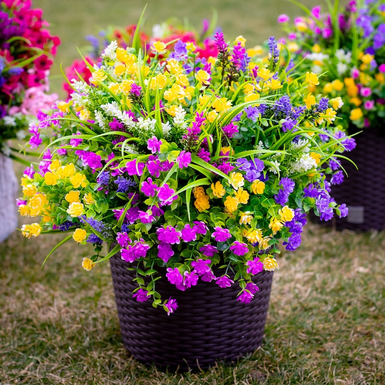 May include: A vibrant arrangement of artificial flowers in a dark brown woven planter. The colorful mix includes yellow, purple, pink, and white blossoms with green foliage. The planter sits on a grassy surface, showcasing a variety of colorful blooms.