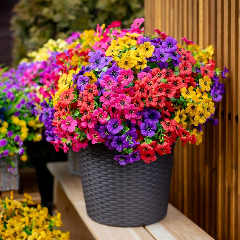 May include: A large, overflowing pot of colorful flowers in shades of red, pink, purple, yellow, and magenta. The flowers are in full bloom and are contained in a dark gray woven-textured pot. The pot sits on a wooden surface.