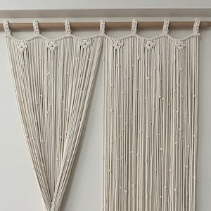 May include: A pair of white macrame curtains with a beaded design. The curtains are hung on a wooden rod and are made of natural fibres.