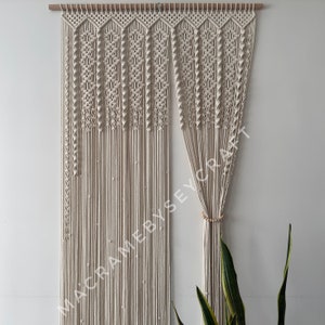 May include: A white macrame curtain with a wooden dowel rod at the top. The curtain is made of a series of knotted cords that create a geometric pattern. The curtain is hanging on a white wall. The text "MACRAME BY SEYCRAFT" is visible in the lower left corner of the image.