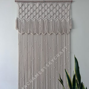 以下が含まれることがあります： 上部に幾何学模様、下部に結び目のついた紐のフリンジが付いた、大きなオフホワイトの糸で編まれた壁掛け。壁掛けは小さな白いビーズで飾られています。