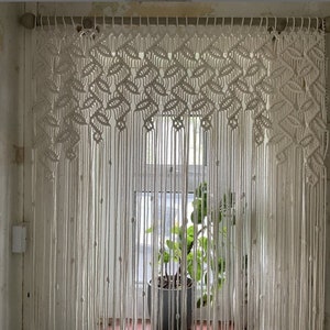 Pode incluir: Cortina de macramé branca com um padrão de folhas pendurado em uma janela. A cortina é feita de cordão de algodão e tem um estilo natural e boêmio.