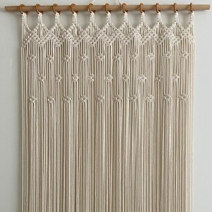 May include: A white macrame wall hanging with a geometric pattern. The hanging is made of cotton cord and is attached to a wooden dowel rod with wooden rings.